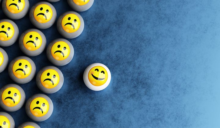Collection of white balls with sad emoji faces on them with a single happy face sitting separately.